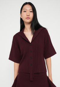 Top de malha canelada bordô com um colarinho largo, mangas curtas, fecho de botões à frente e um detalhe de laço na bainha, apresentando um acabamento texturizado.