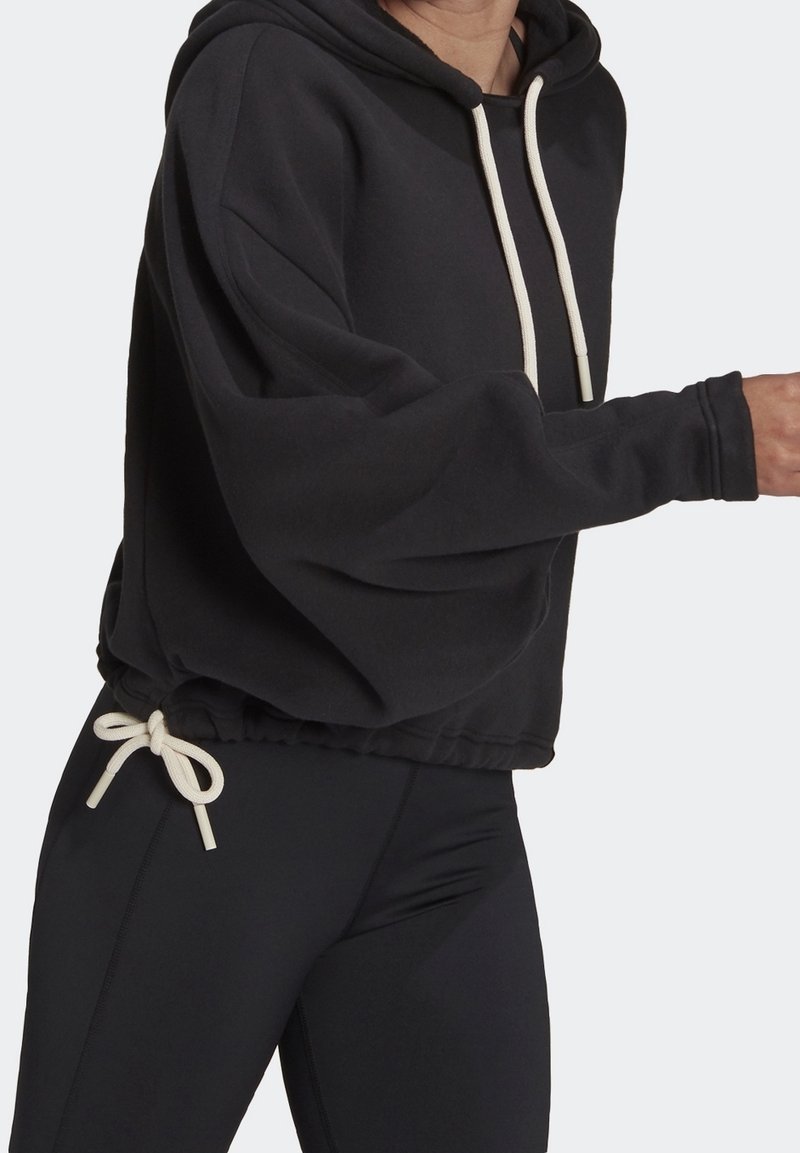 Personne portant un sweat à capuche noir avec des cordons blancs et des leggings noirs, partiellement visible du cou jusqu'à mi-cuisse.