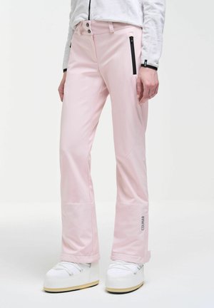 Personne portant un pantalon de ski rose clair avec des poches zippées noires et des chaussures de ski blanches, debout devant un fond uni.