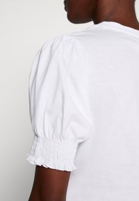 Primo piano del braccio superiore di una persona di pelle scura che indossa una camicia bianca con maniche a palloncino e polsini elasticizzati arricciati.