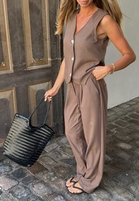 Top marrone senza maniche con bottoni, pantaloni larghi coordinati e una borsa a tracolla nera intrecciata. Pietre naturali sotto i piedi.