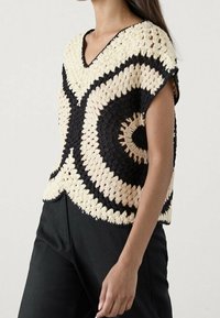 Crochet-huppari, jossa on V-niskamuotoilu, musta-kermanvärisiä pyöreitä kuviota, lyhyet hihat ja löysä istuvuus.