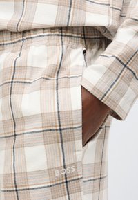 Mäkké kárované oblečenie na doma v béžovej, bielej a čiernej farbe. Elastický pás s bočnými vreckami, zvýraznený diskrétnym detailom loga.