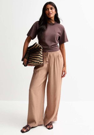 T-shirt en coton marron, pantalon large beige avec taille à cordon de serrage et sac fourre-tout tissé noir, avec un motif en zigzag texturé. Sandales noires.