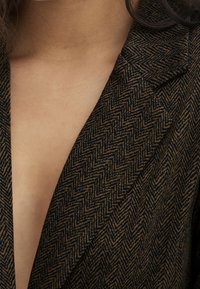 Gros plan sur la poitrine portant un blazer marron et noir à motif chevrons avec un décolleté en V profond.