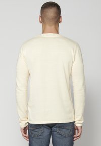 Helles Creme-farbenes Langarmshirt mit strukturierten Rippärmel und glattem Körper. Die Rückansicht zeigt einen klassischen Rundhalsausschnitt und eine figurbetonte Silhouette.