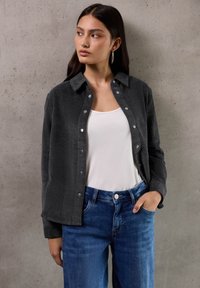 Grijze corduroy button-up shirt met een kraag, gecombineerd met een witte tanktop en blauwe jeans. Model gepresenteerd tegen een neutrale achtergrond.