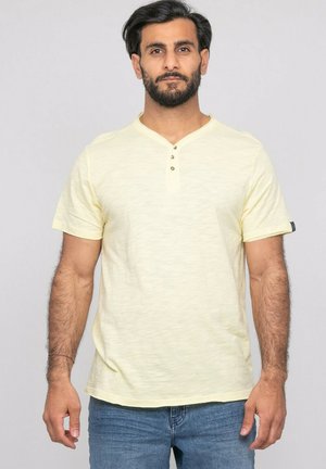 Homme barbu portant un t-shirt henley jaune pâle à manches courtes et un jean bleu, debout devant un fond uni gris clair.