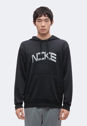 Joven con sudadera negra con capucha con texto blanco "NIKE" y pantalones grises, de pie mirando hacia adelante contra un fondo claro y liso.