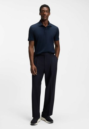 Mand, der står iført en mørkeblå tekstureret kortærmet polo, sorte plisserede bukser og sorte sneakers med hvide såler.
