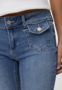 Blå denimjeans med en åtsittande design, med två framfickor och en bakficka med ett distinkt sömmönster. Metallknappstängning.