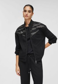 Veste noire zippée avec un design texturé ornée de strass sur les épaules. Comprend des poignets côtelés et une coupe décontractée.