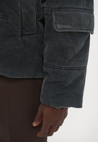 Musthave mustra nahk jakk, millel on tekstureeritud pind, küljetasku ja tikandite detailid. Varruka serv on veidi rullitud.