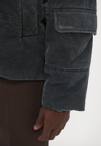 Sort fløjlskjorte med en struktureret overflade, der har en sidelomme og broderidetaljer. Måtten er let rullet op.
