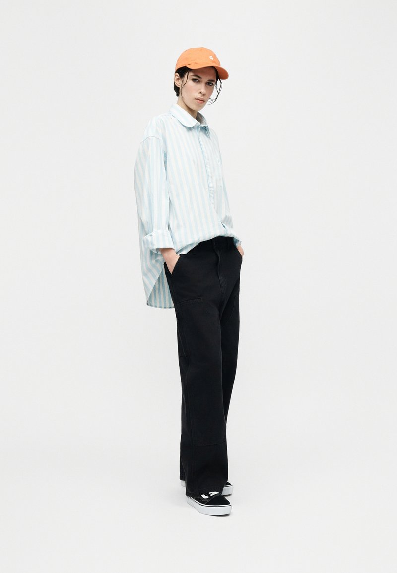 Giovane con indosso una camicia rigata azzurro chiaro oversize, pantaloni larghi neri, cappellino arancione e sneakers nere, in piedi con le mani nelle tasche.