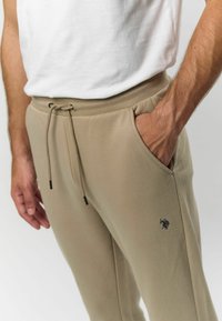 Béžové tepláky vyrobené z mäkkého materiálu, s elastickým pásom na šnúrku, bočnými vreckami a jemným logom na stehne.