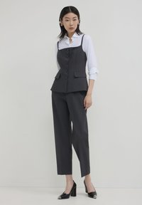 Donna che indossa un gilet nero con bottoni sopra una camicia bianca, pantaloni neri su misura e tacchi neri con punte metalliche, in piedi contro uno sfondo chiaro e uniforme.