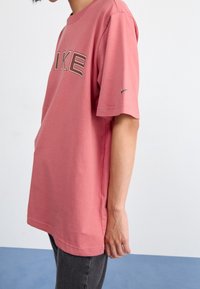 Růžové bavlněné tričko s krátkými rukávy, se znakem "NIKE" v tmavě šedé barvě a malým Nike swoosh na levém rukávu.