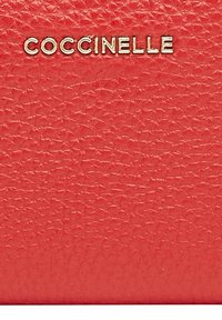 Rød teksturert skinnlommebok med en gull "Coccinelle"-logo. Har en småsteinsoverflate og kompakt rektangulær form.