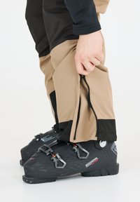 Pantaloni da sci beige e neri, con cerniere alle caviglie, abbinati a scarponi da sci neri con accenti rossi e argentati.