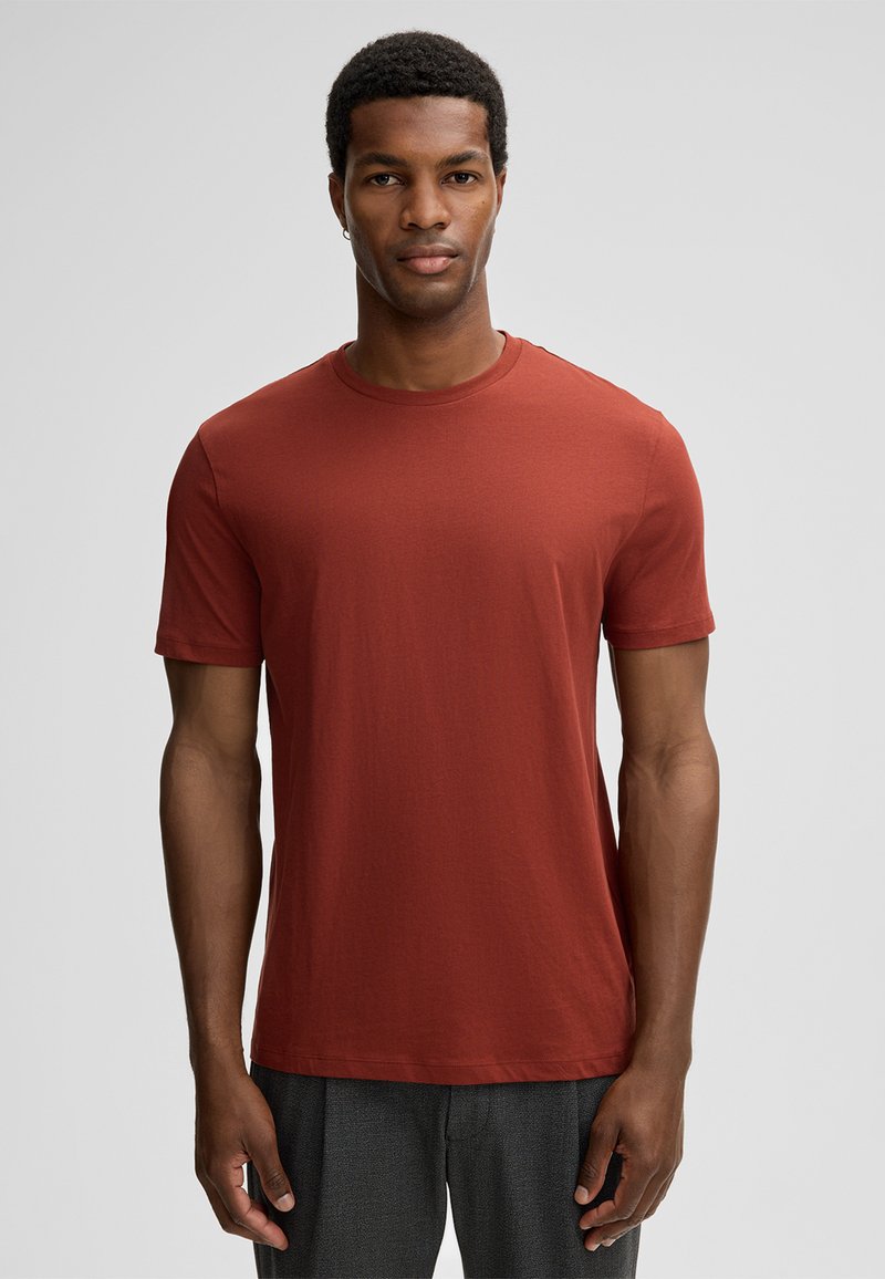 Man staat rechtop en kijkt naar voren, gekleed in een effen roestkleurig T-shirt met korte mouwen en donkergrijze broek, tegen een lichtgrijze achtergrond.