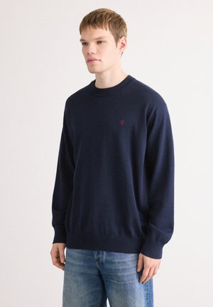Marineblauwe gebreide trui met een ronde halslijn en lange mouwen. Heeft een klein rood logo op de borst en ribgebreide manchetten en onderkant.
