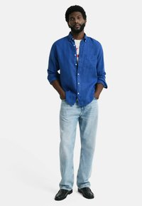 Camisa azul de botones con un bolsillo en el pecho, combinada con jeans de un azul claro y zapatos negros. La camisa tiene las mangas arremangadas y un corte relajado.