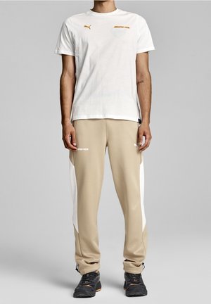 Osoba w białej koszulce Puma AMG, beżowych spodniach dresowych Puma AMG z białymi panelami po bokach oraz czarnych butach sportowych, stojąca na tle szarego tła.