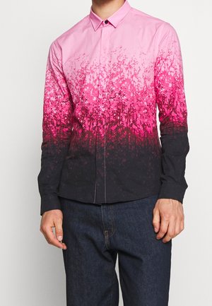 Mann trägt langärmeliges Hemd mit Farbverlauf von Pink zu Schwarz und dunkle Jeans, steht vor einfachem Hintergrund.