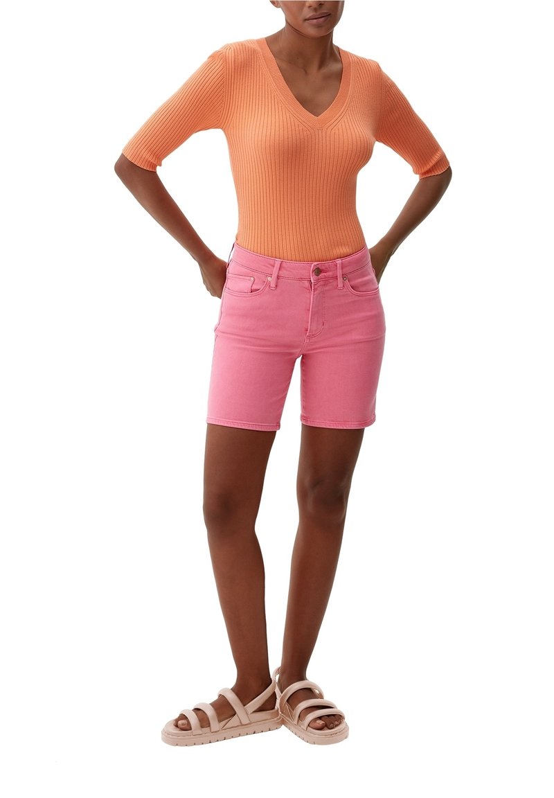 Mujer vistiendo una blusa naranja de punto con escote en V, pantalones cortos de mezclilla rosa y sandalias beige, de pie con las manos en las caderas contra un fondo blanco.