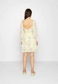 Vestido curto amarelo com padrão floral, mangas longas, costas abertas e punhos com frufrus. Combinado com sapatos de plataforma verdes.