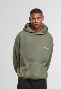 Joven con una sudadera de algodón verde oliva de gran tamaño que tiene un bolsillo frontal y el texto "Sunday Running Club" en el pecho, de pie contra un fondo liso.