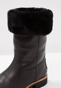 Botte en cuir noire mi-mollet avec bordure en fourrure noire épaisse et semelle cousue marron, conçue pour l'hiver.