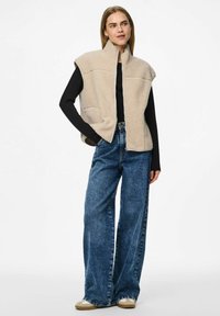 Vrouw in een beige fleecvest over een zwart langemouwen top, met wijd uitlopende blauwe jeans en beige sneakers, staande met één hand in de zak.