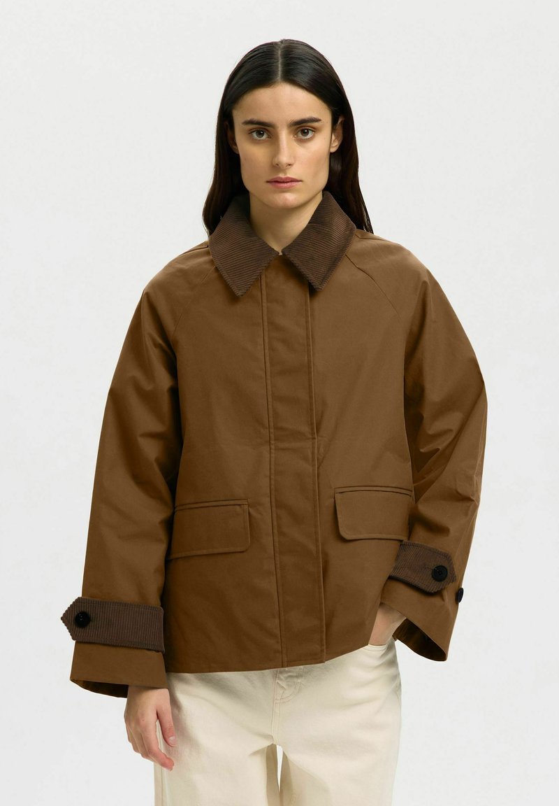 Femme aux longs cheveux foncés portant une veste marron avec col et poignets en velours côtelé, posant devant un fond clair uni.