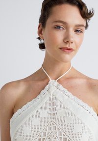 Top bianco con scollo all'americana caratterizzato da un bordo in pizzo testurizzato e modelli geometrici all'uncinetto, dotato di un laccio al collo e un design con scollo aperto.