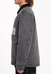 Veste en toile cirée grise avec un col en velours côtelé noir, poche avant avec un patch décoratif et des détails de couture subtils.