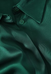 Mörkgrön satin skjorta med spetsig krage. Har två matchande knappar och en slät textur som visar en lätt glans och drapering.