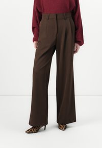 Braune weit geschnittene Hose aus glattem Stoff mit Falten. Kombiniert mit einem roten Oversized-Pullover und hochhackigen Schuhen im Animal-Print.