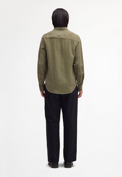 Osoba stojąca tyłem, ubrana w oliwkowozieloną koszulę z długim rękawem, czarne spodnie i czarne buty na jednolitym białym tle.