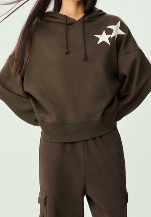 Woman wearing a dark brown cropped hoodie with two white star patches on shoulder and matching cargo pants against light background.