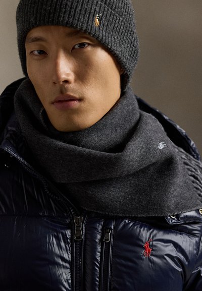 Temno siva pletena kapa in šal v kombinaciji z mornarsko modro puhovko. Obleka ima vidne logotipe in gladko, sijočo teksturo na jakni.