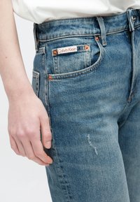 Mano descansando junto a unos vaqueros de mezclilla azul claro con una pequeña etiqueta de Calvin Klein cerca del bolsillo delantero y un sutil detalle de desgaste en la tela.