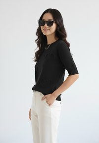 Czarny dziany sweter z krótkimi rękawami, ozdobiony teksturowanymi wzorami serc, w połączeniu z białymi spodniami i ciemnymi okularami przeciwsłonecznymi.