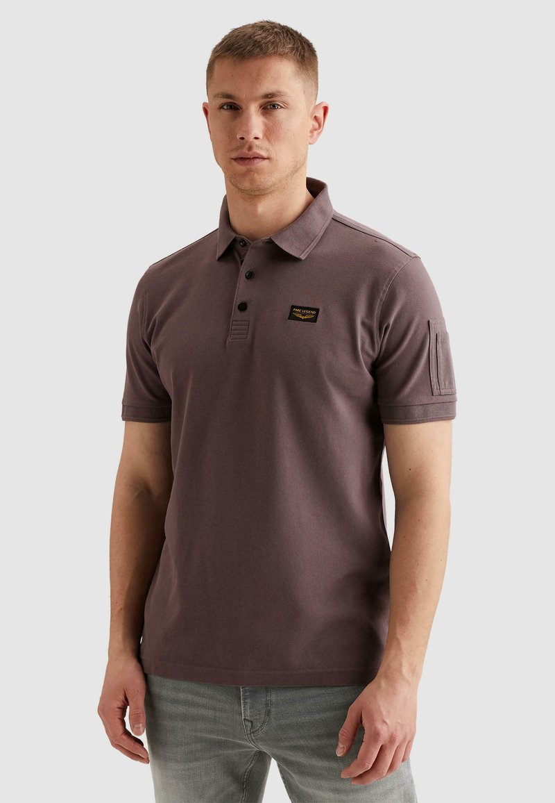 Homme portant un polo marron à manches courtes avec un col boutonné et un écusson sur la poitrine, associé à un jean gris clair, debout de manière neutre.