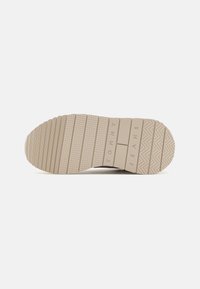 Suela de zapato de goma beige con surcos texturizados, líneas horizontales y verticales, y el logo emblema 'TOMMY JEANS' en relieve.