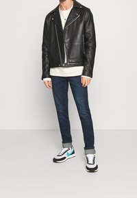 Svart lädermotorcykeljacka med asymmetrisk dragkedja, vit t-shirt med rund hals, mörkblå jeans och svarta och vita sneakers med turkosa detaljer.