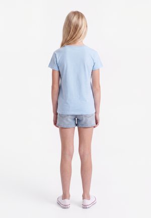 Barn med langt blondt hår står med ryggen til iført en lyseblå t-shirt, denimshorts og hvide sneakers på en hvid baggrund.