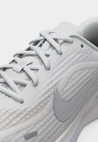 Κοντινή λήψη λευκού παπουτσιού για τρέξιμο Nike με υφασμένο πάνω μέρος με υφή, λογότυπο Swoosh σε ανοιχτό γκρι και λεπτομερείς λευκές κορδόνια που περνούν μέσα από θήκες.