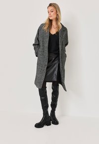 Grijze textuurjas met een oversized pasvorm, zwarte top, leren rok en zwart leren kniehoog hakken. Eenvoudig, gestructureerd ontwerp met zakken.
